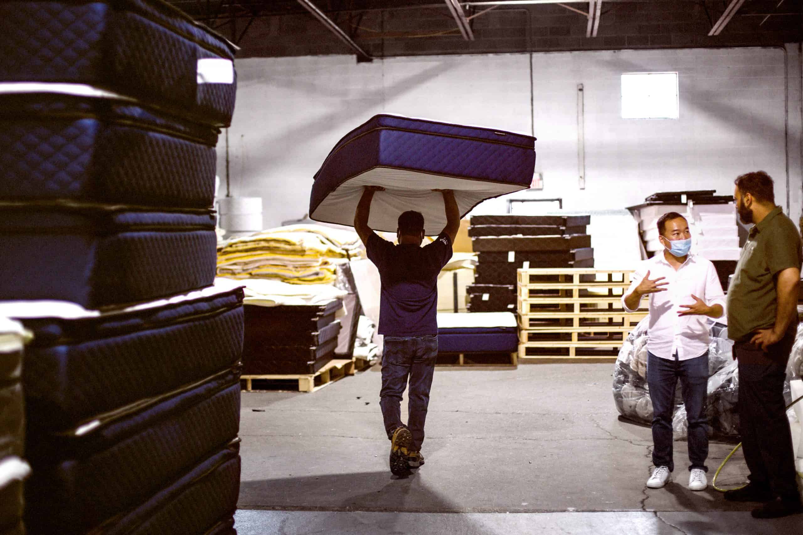 A man walks with a Silk & Snow mattress over his head as two other men have a conversation.